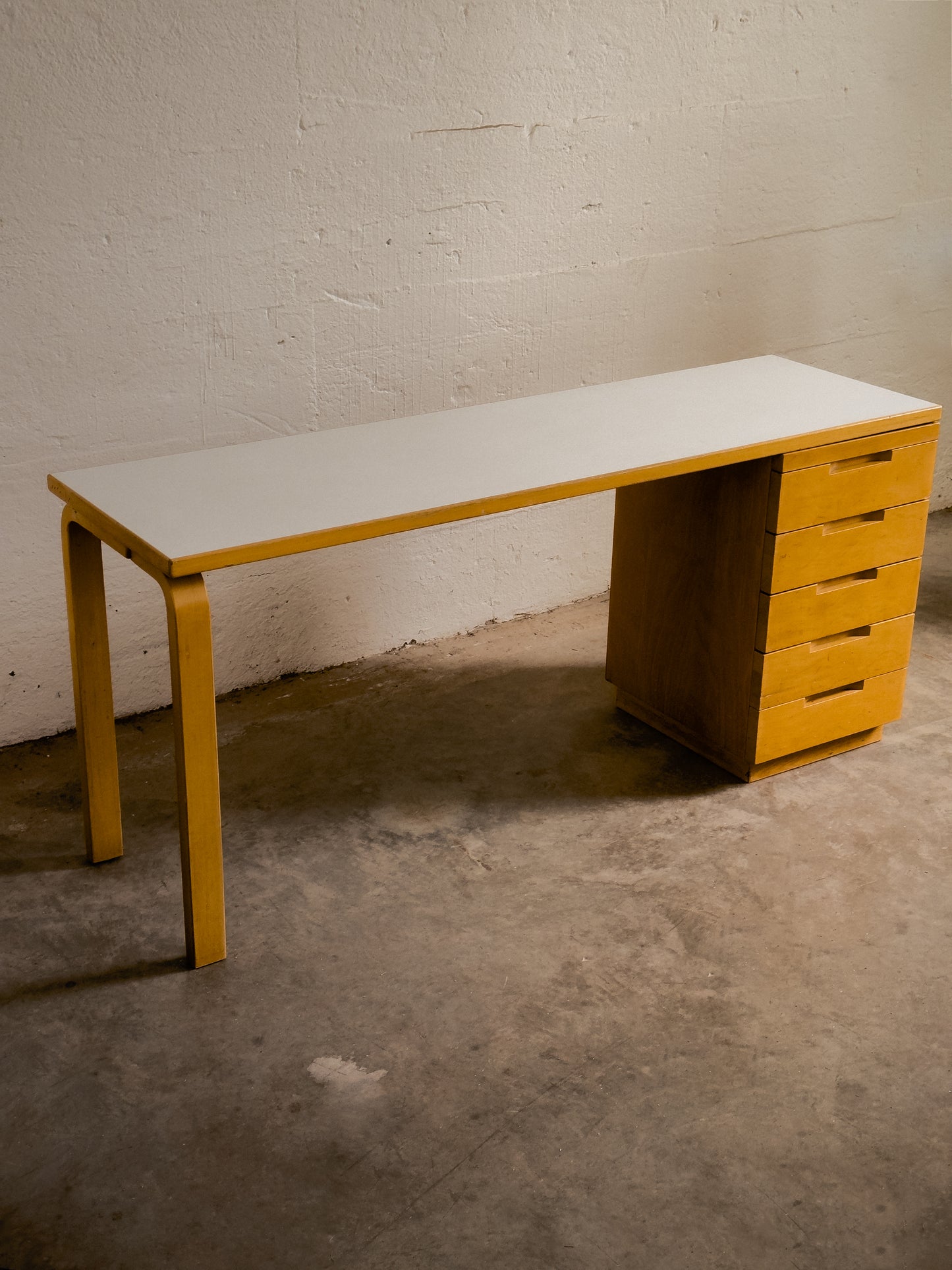 Aalto Wooden desk with drawers and chair against a white brick wall.