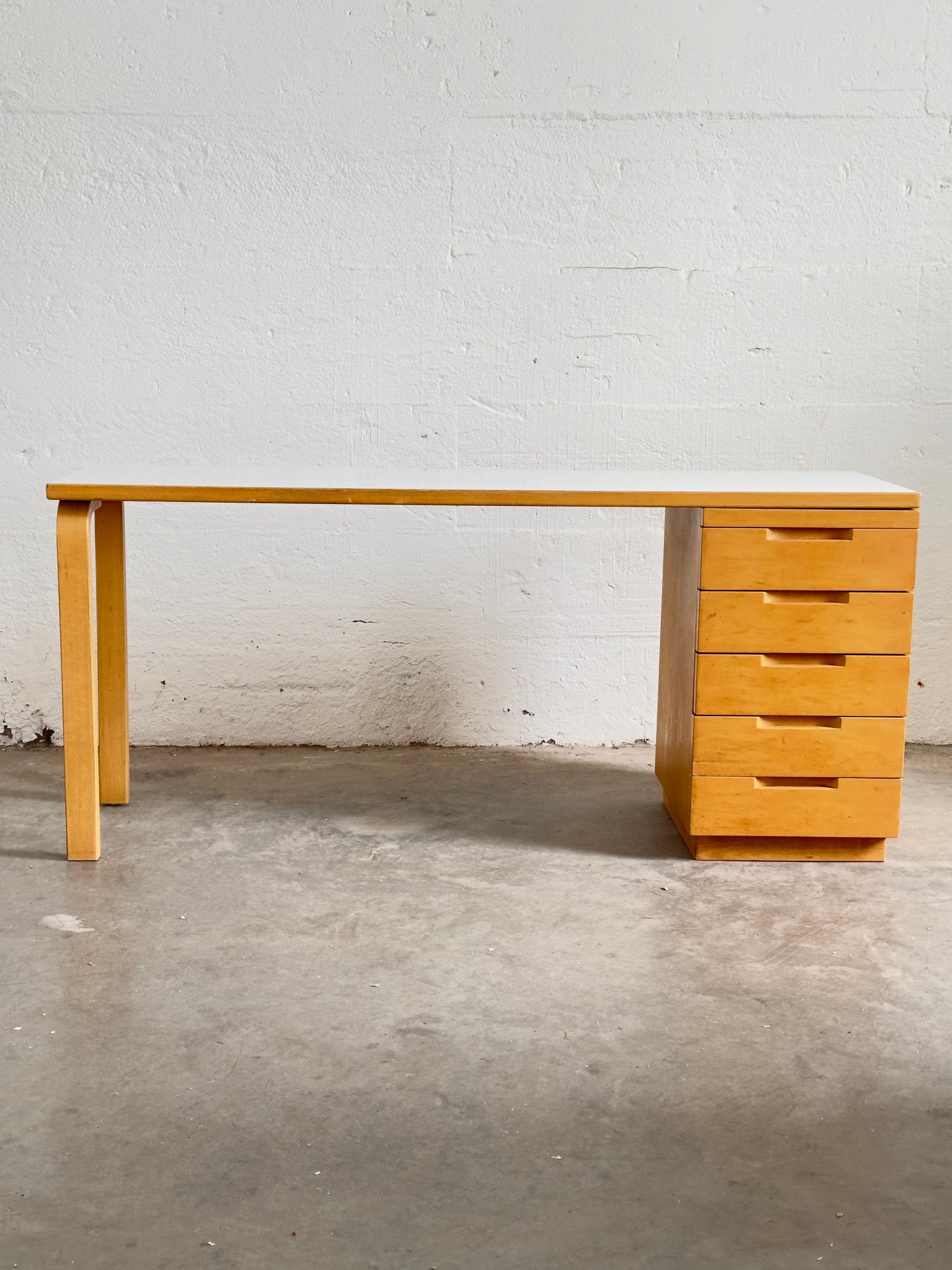 Aalto Wooden desk with drawers and chair against a white brick wall.